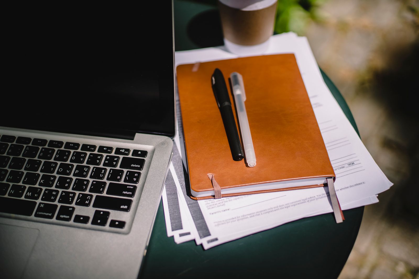 A notebook, pens and printed forms stacked beside a laptop on a desk