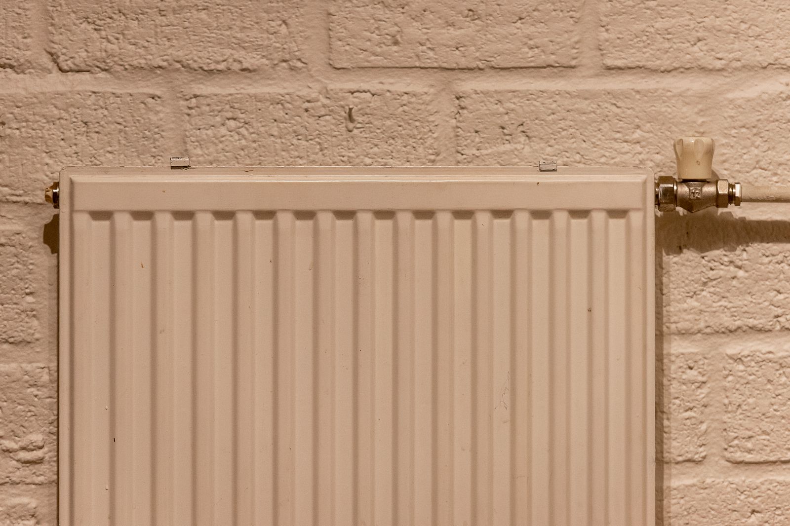 A white panel radiator with a valve at one end, fitted against a painted brick wall